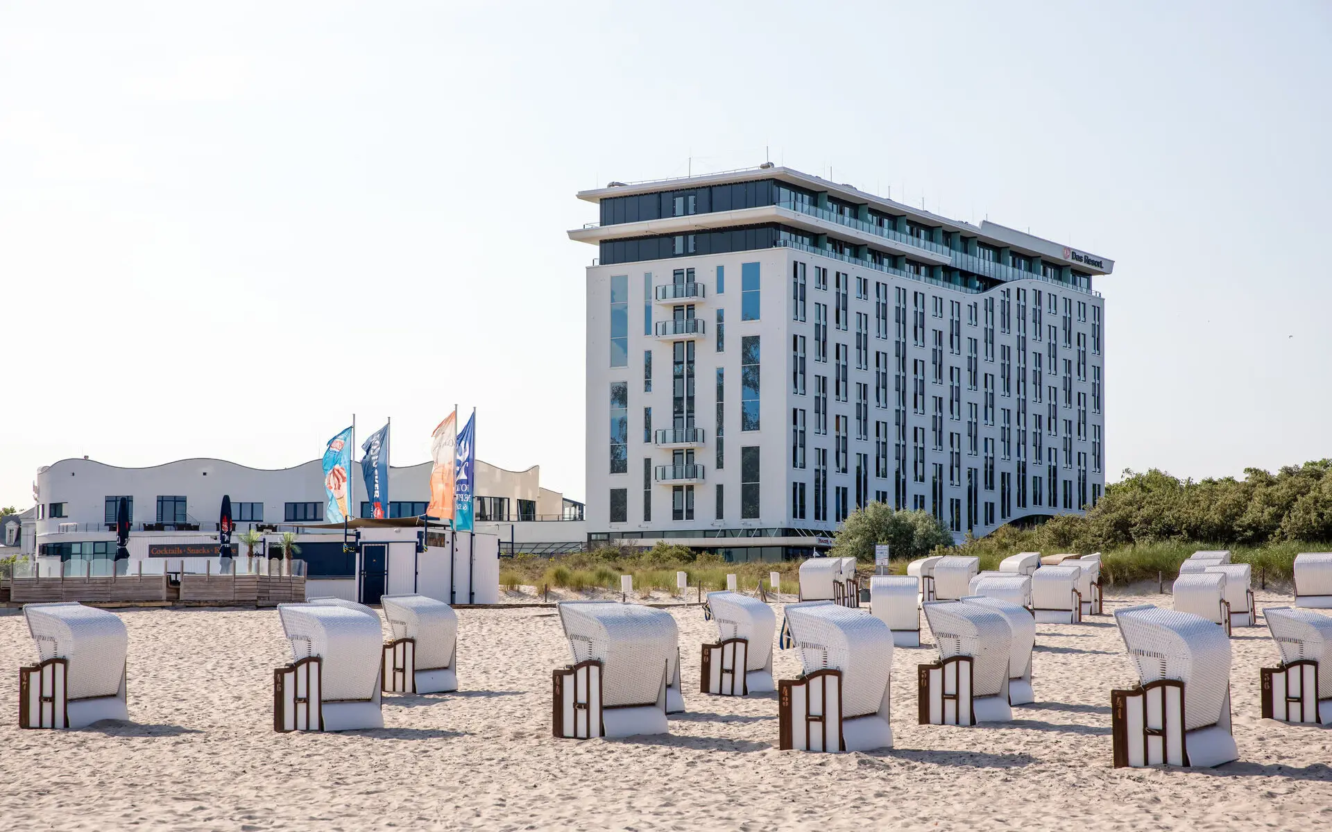 Eine Außenansicht des aja Warnemünde als weißes Gebäude mit vielen Fenstern an einem Strand mit weißen Strandkörben.