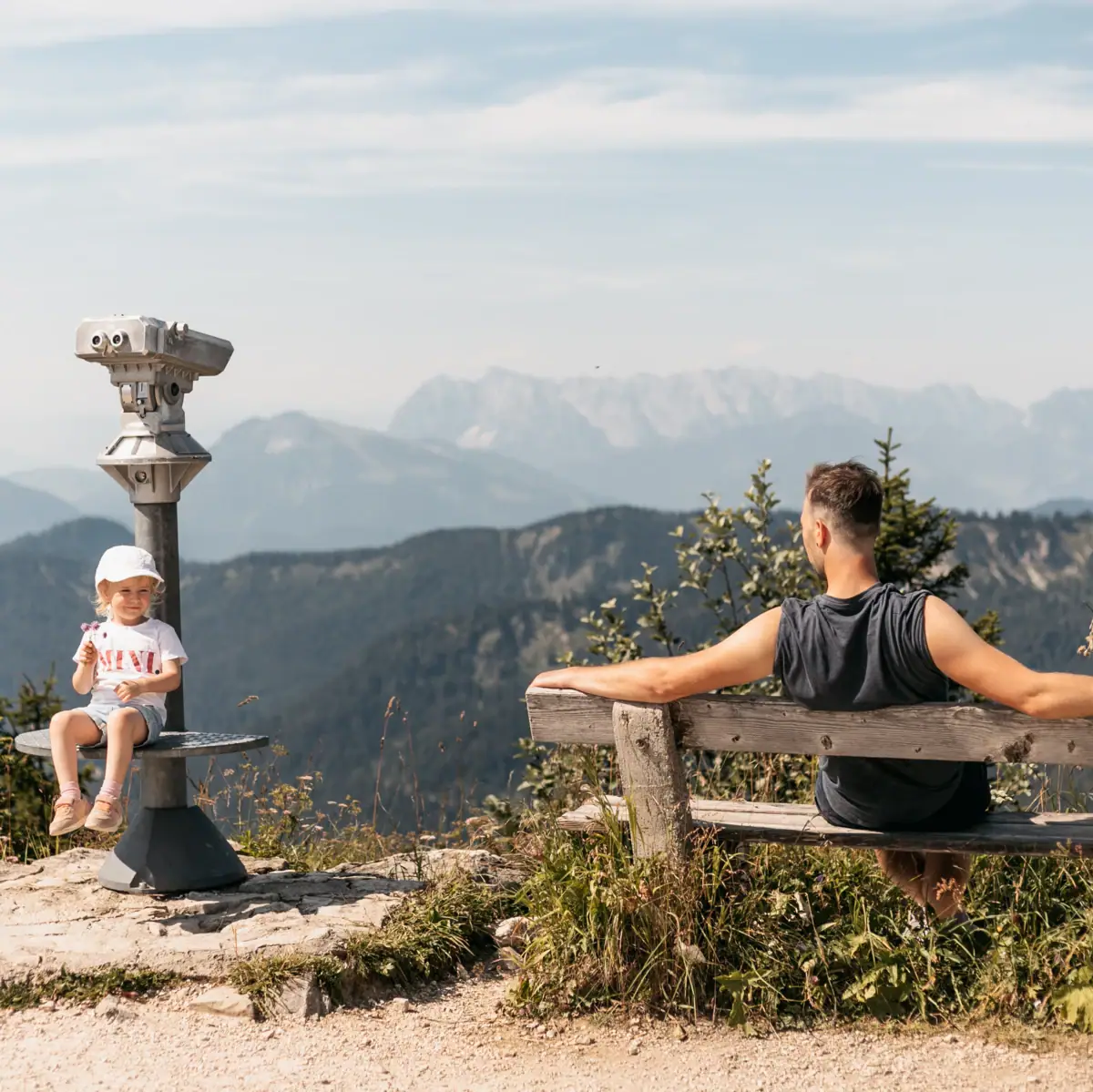 Ein Mann und ein Kind sitzen auf einer Bank mit Blick auf Berge.