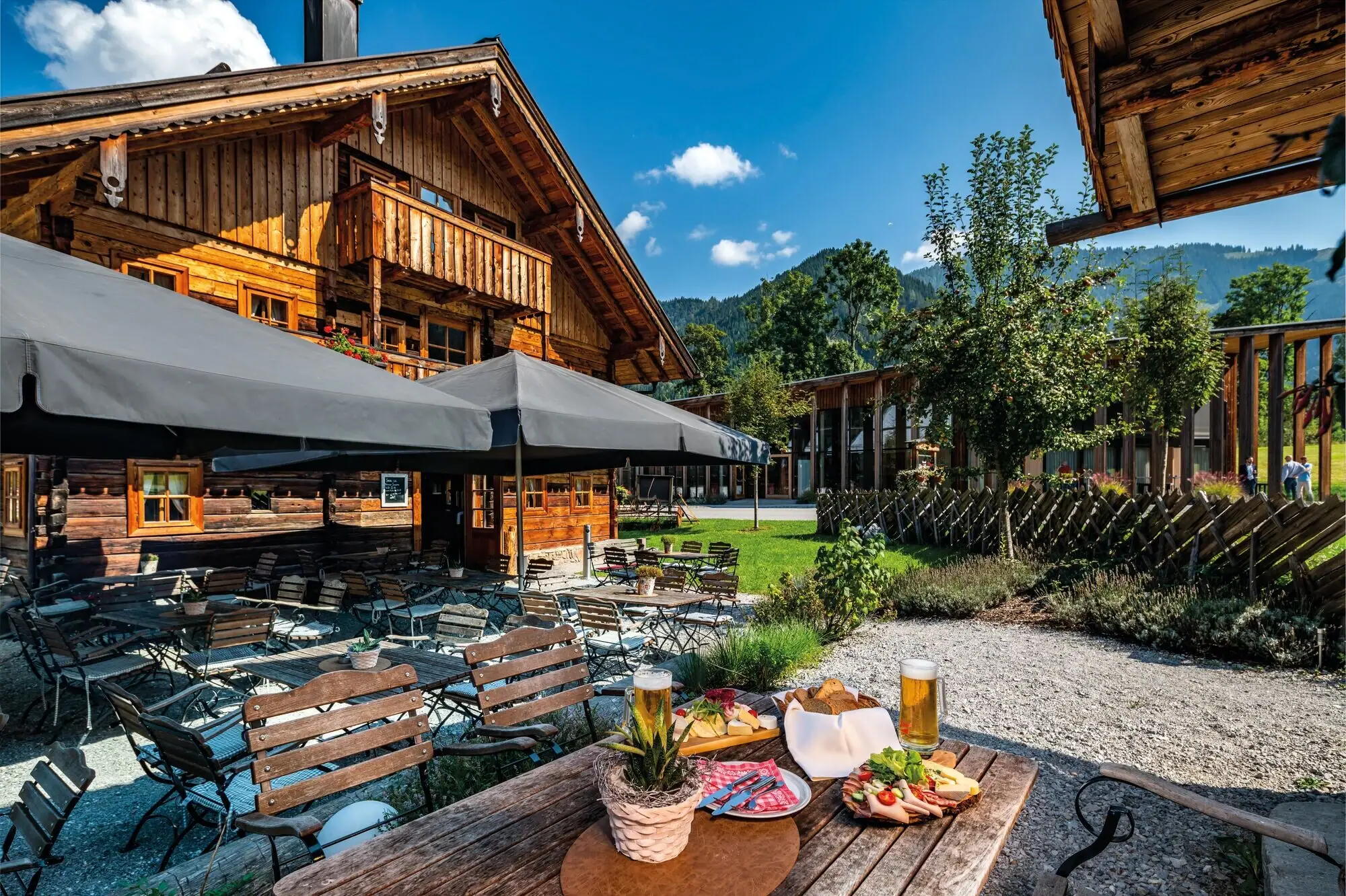 Gemütlicher Biergarten vor einer traditionellen alpinen Holzhütte mit rustikalem Charme in Werfenweng. Holztische und -stühle unter großen Sonnenschirmen laden zum Verweilen ein. Im Vordergrund stehen ein Bierglas, eine Brotzeitplatte mit Wurst, Käse und frischem Gemüse sowie ein gedeckter Tisch.