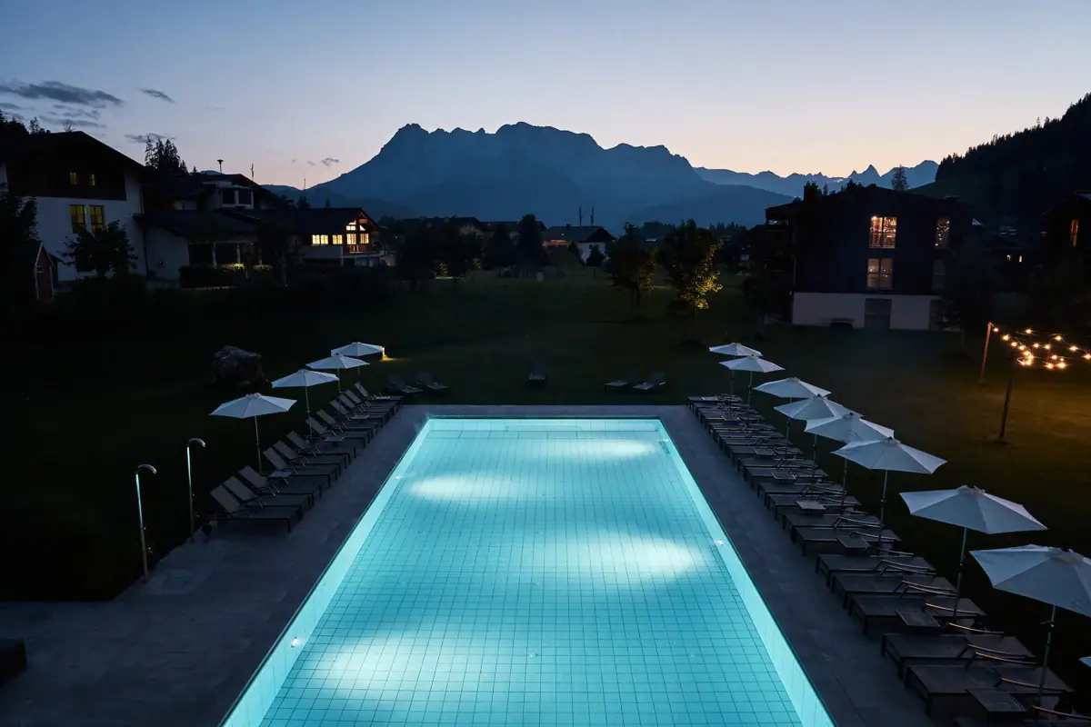 Ein Pool mit Stühlen und Sonnenschirmen direkt am Hotel. Am Horizont die Berge im Abendlicht.