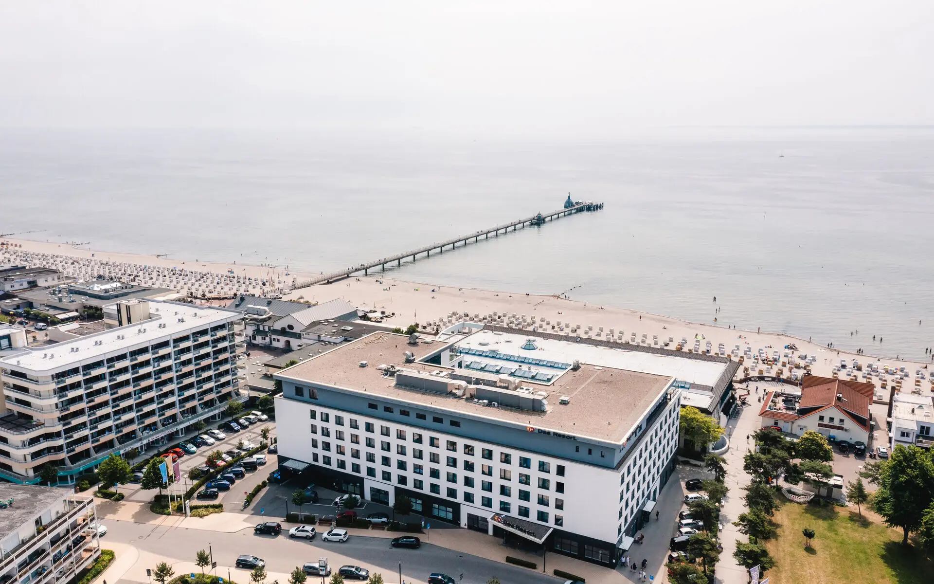 Eine Luftansicht von einem großen Gebäude und weiteren umliegenden Gebäuden am Strand, von welchem aus ein langer Steg in das angrenzende Meer führt. 