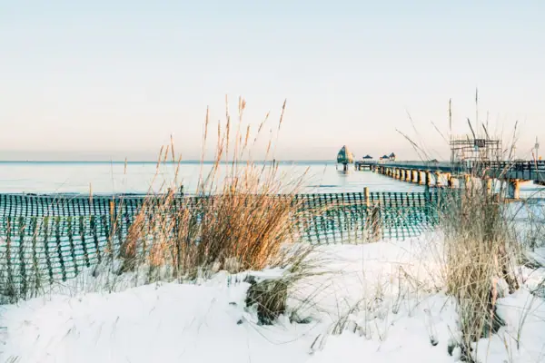 Schneebedeckter Strand mit Steg und Gräsern im Winter.