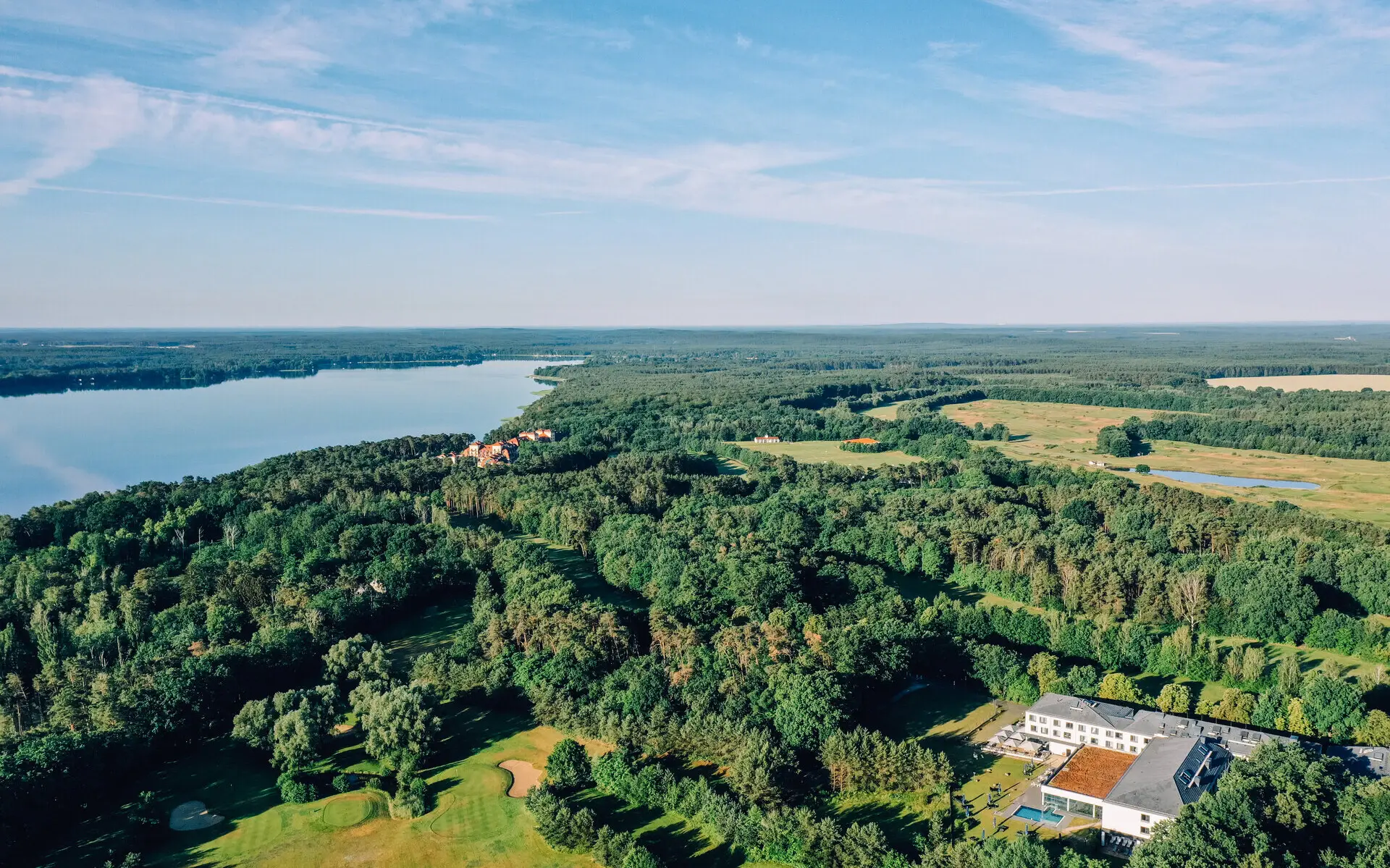 Die Luftansicht einer grünen Landschaft auf der ein weißes Gebäude steht, umgeben von Wäldern und einem großen See im Hintergrund. 