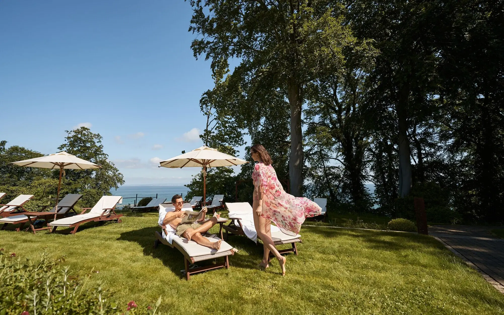 Paar entspannt auf Sonnenliegen im Garten eines Hotels mit Ostseeblick; die Frau trägt ein luftiges Sommerkleid, der Mann prostet mit einem Getränk – Sonnenschirme und grüne Bäume im Hintergrund.
