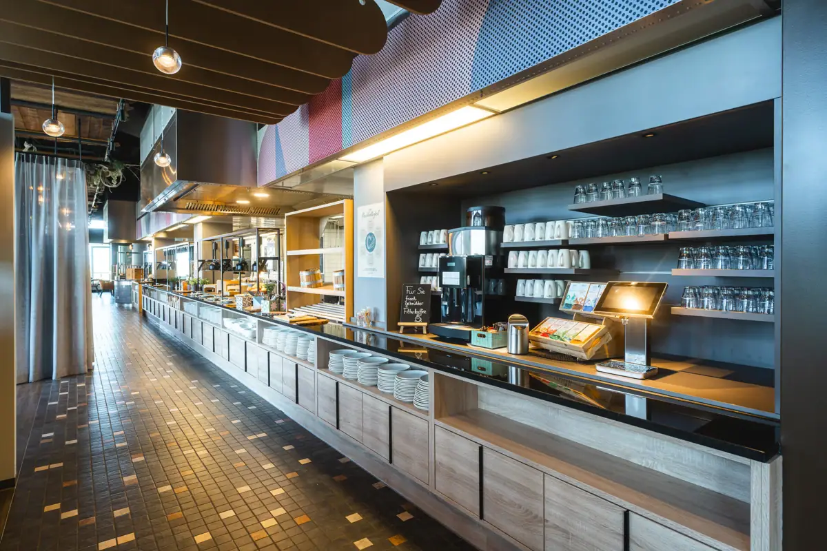 Restaurant im aja Warnemünde Das Buffetrestaurant mit Tresen und Regalen voller Geschirr.