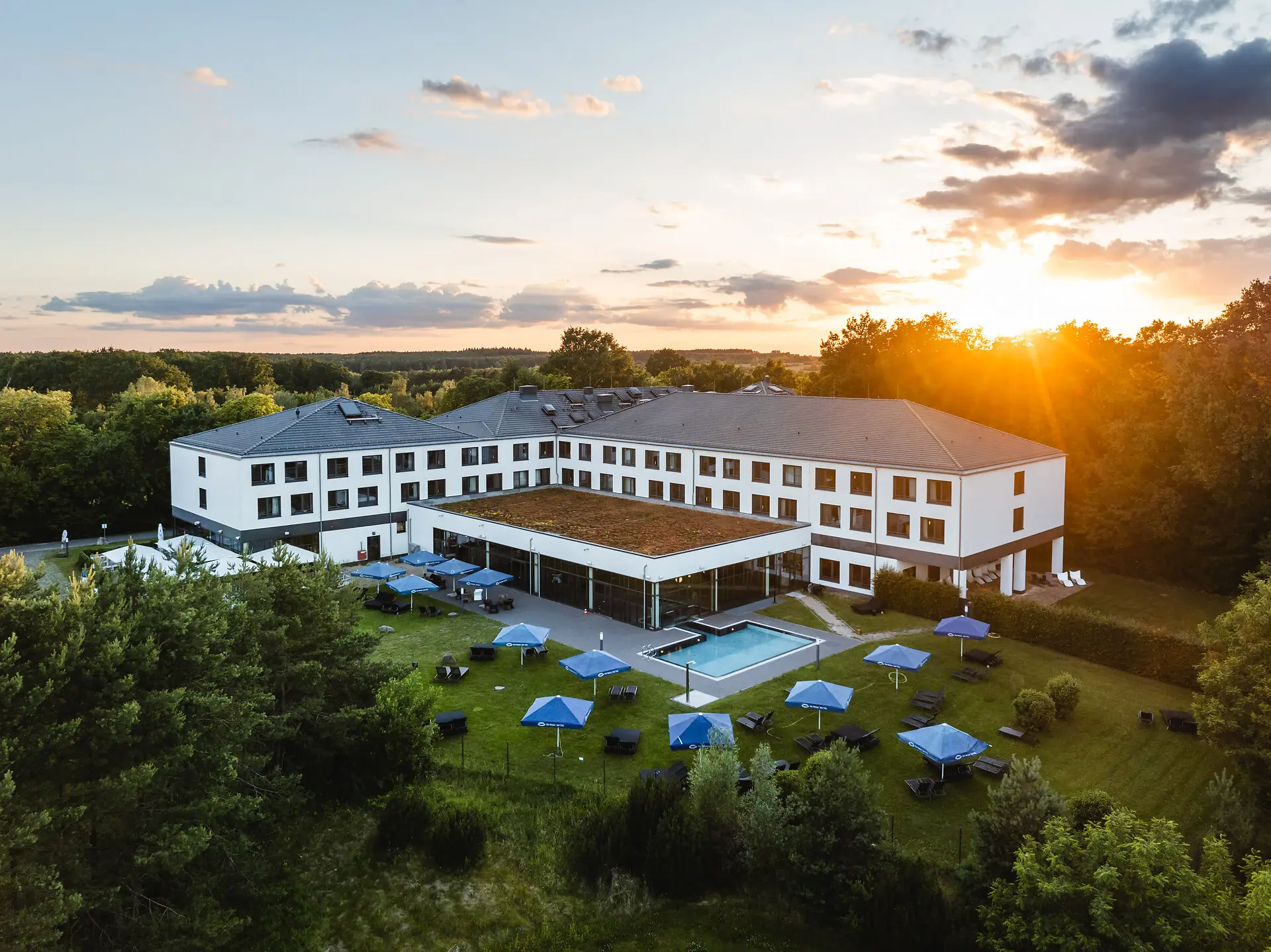 Die Außenansicht des aja Bad Saarow mit Außenpool und blauen Sonnenschirmen auf der Wiese. Die umliegenden Wälder sind grün und die Sonne geht über den Bäumen auf. 