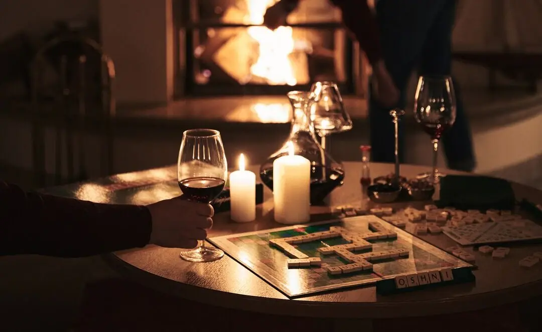 Im Vordergrund steht ein Tisch mit einem Gesellschaftsspiel, brennenden Kerzen, einem Glas Rotwein und Snacks. Im Hintergrund kümmert sich eine Person um das Feuer im Kamin, umgeben von stimmungsvoll arrangierten Kerzen auf dem Kaminsims