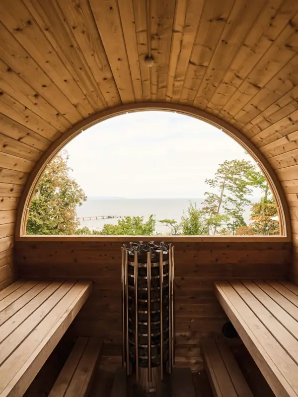Eine Fasssauna aus Holz mit rundem Fenster, das auf das Wasser blickt.