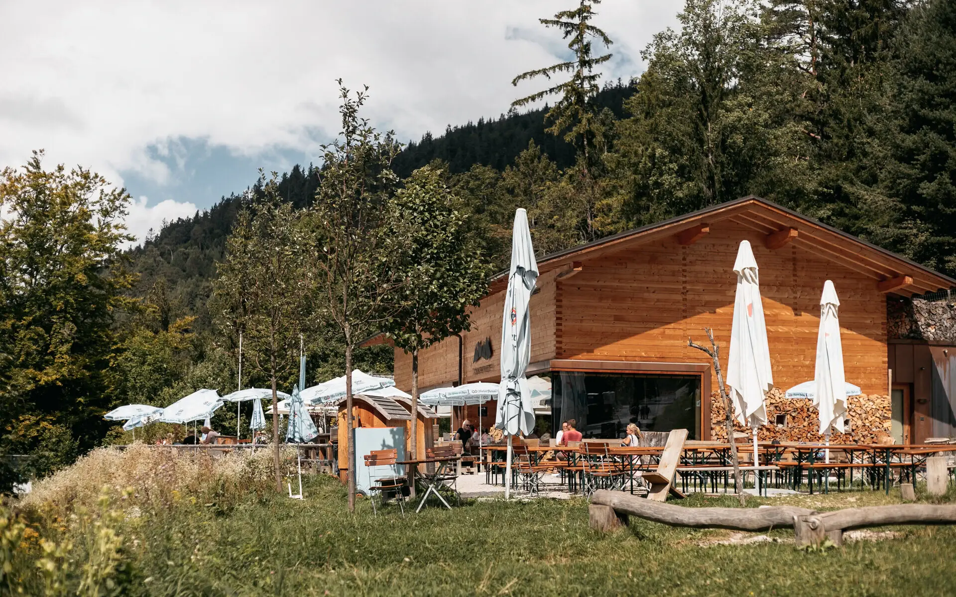 Eine gemütliche Alm in Ruhpolding mit einer sonnigen Terrasse, bestückt mit Holzbänken und Tischen, umgeben von grüner Natur und dichten Wäldern. 