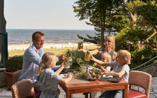 Eine Familie mit drei Kindern sitzt auf einer Terrasse an einem Tisch, auf dem Essen steht und stößt mit Limonaden an. Im Hintergrund ist das Meer und der Strand zu sehen.