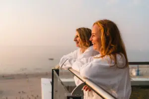 Ausblick Zwei Frauen in Bademänteln lehnen an einem Geländer mit Blick auf einen Strand.