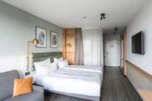 Zimmer im aja Warnemünde Hotelzimmer mit einem Doppelbett, ausgestattet mit Kissen und Bettwäsche. Fernseher an der Wand und Couch neben dem Bett stehend.