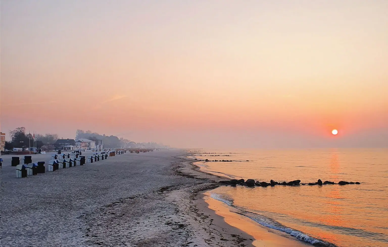 Ein ruhiger Strand an einem Frühlingsmorgen. Tau hängt in der Luft und die warmen Farben der aufgehenden Sonne spiegeln sich im Wasser. 