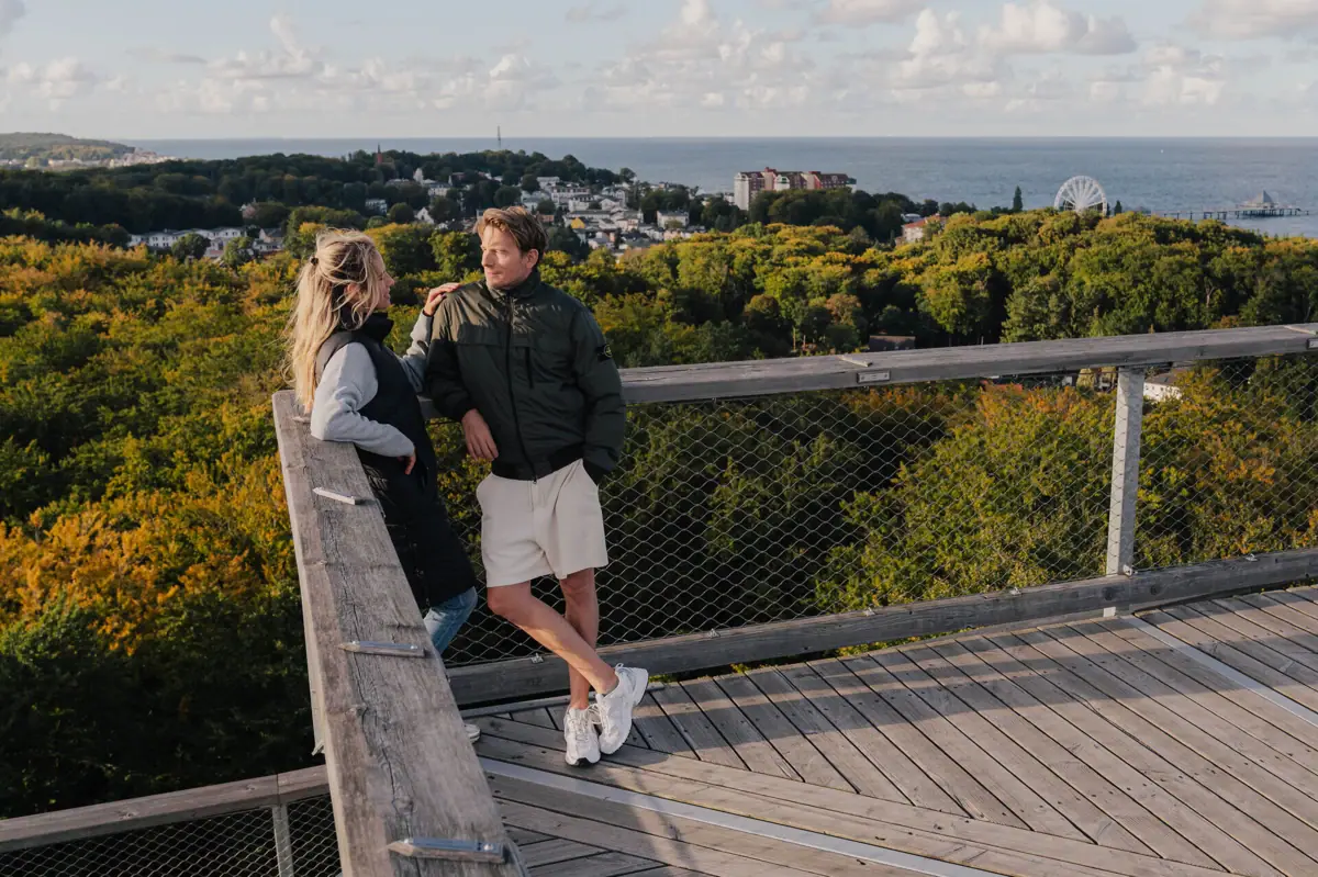 Ein Mann und eine Frau stehen auf einem Holzdeck mit Blick auf einen Wald.