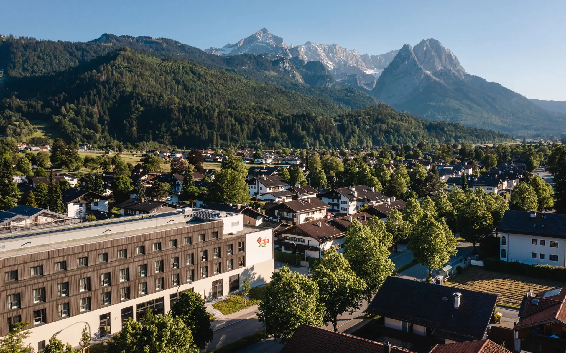 Eine Außenansicht des aja Garmisch-Partenkirchen umgeben von weiteren Häusern der Stadt und im Hintergrund erstrecken sich Wälder und Berge. 