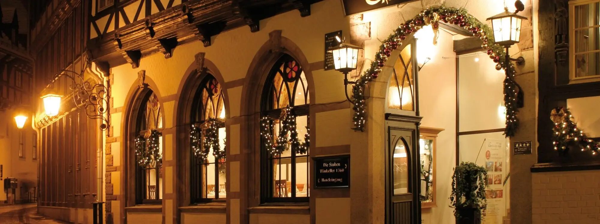 Das festlich beleuchtete Hotel 'Gothisches Haus' bei Nacht, mit geschmückten Fensterbögen und einem einladenden Eingang. Die historische Fachwerkfassade wird von warmem Licht hervorgehoben.