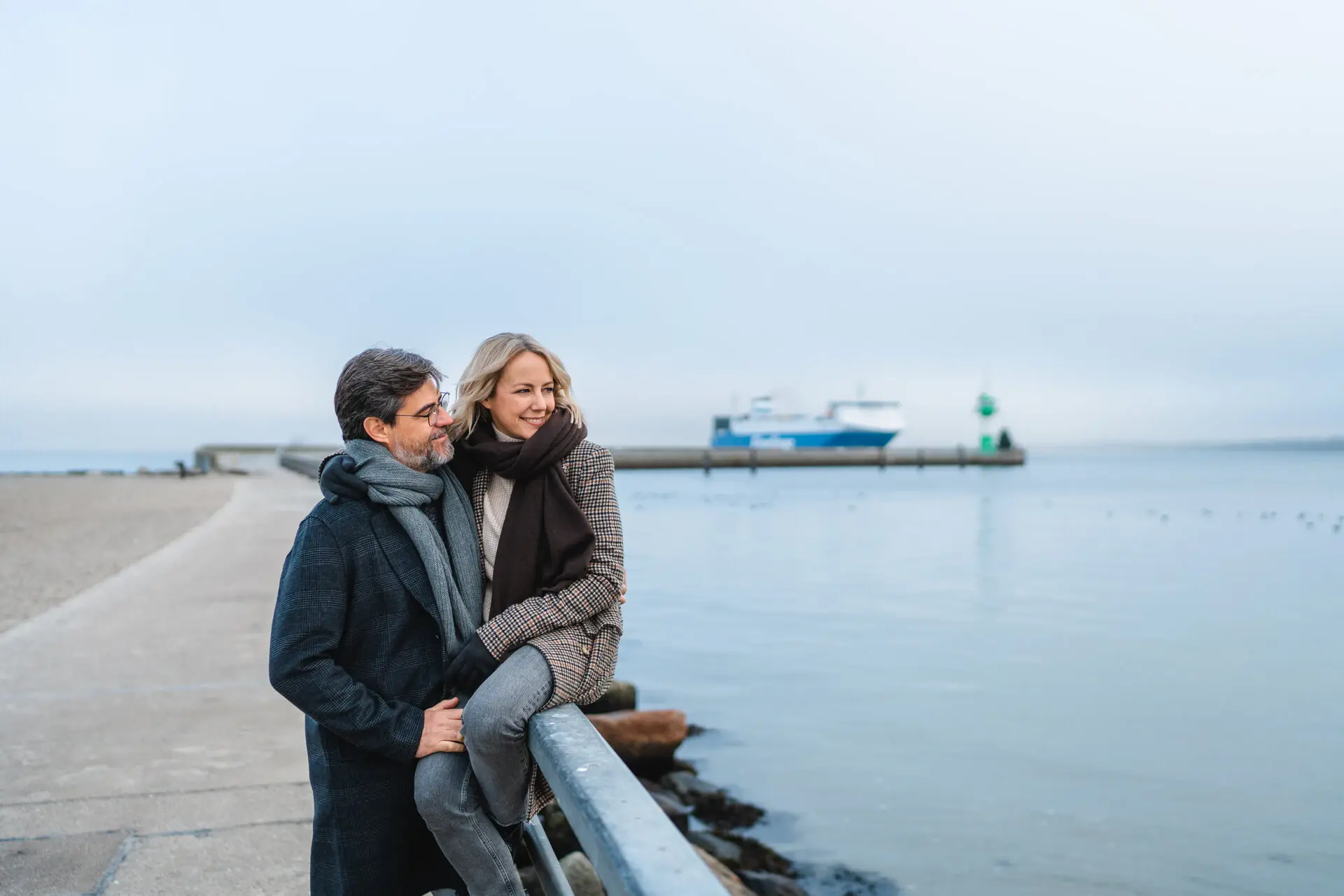 Ein Mann und eine Frau stehen an einem Geländer am Wasser, beide tragen Jeans und Jacken.