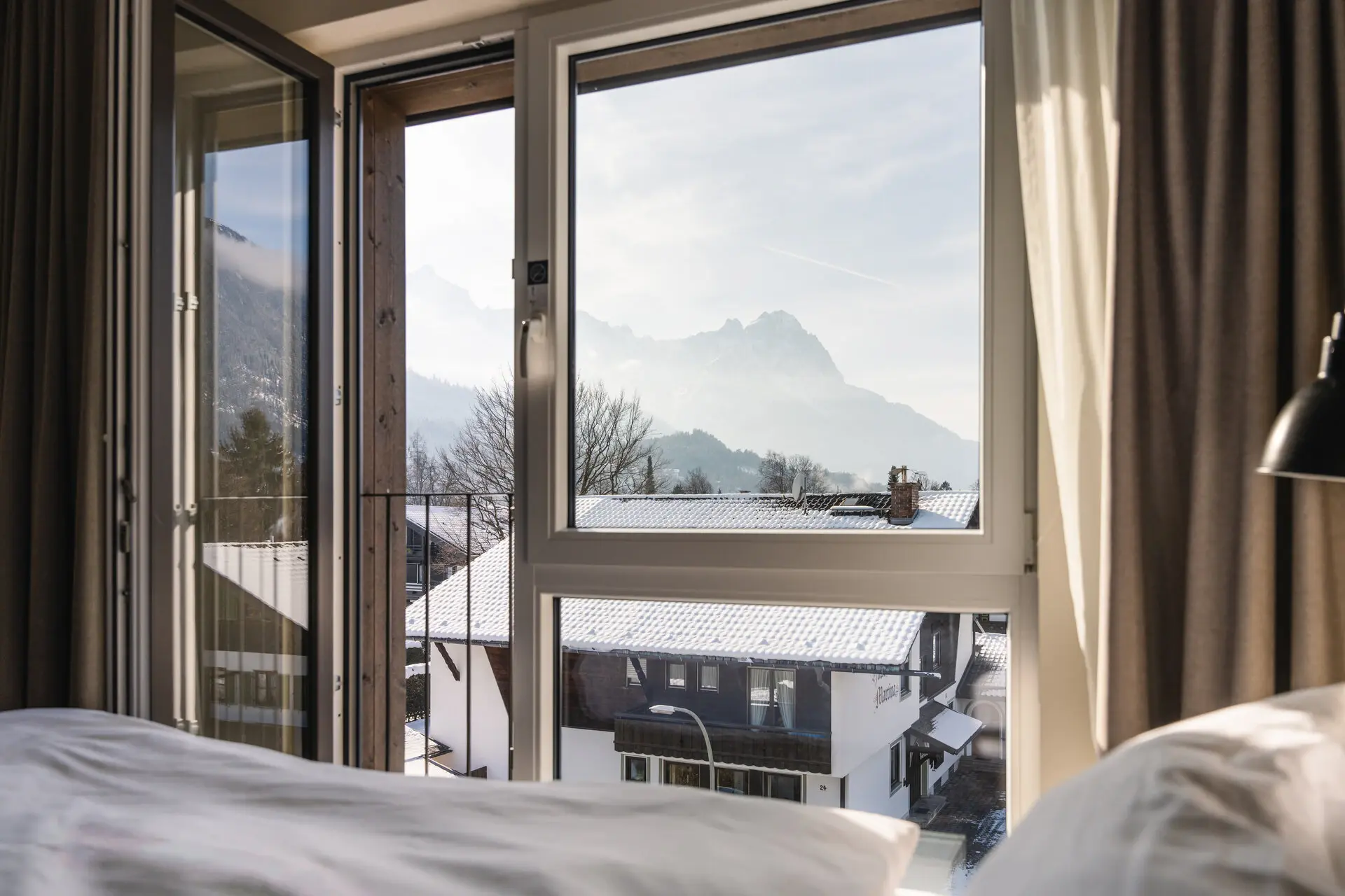 Blick aus einem Fenster auf ein Bergpanorama, mit sichtbarem Bett und Kissen im Vordergrund.