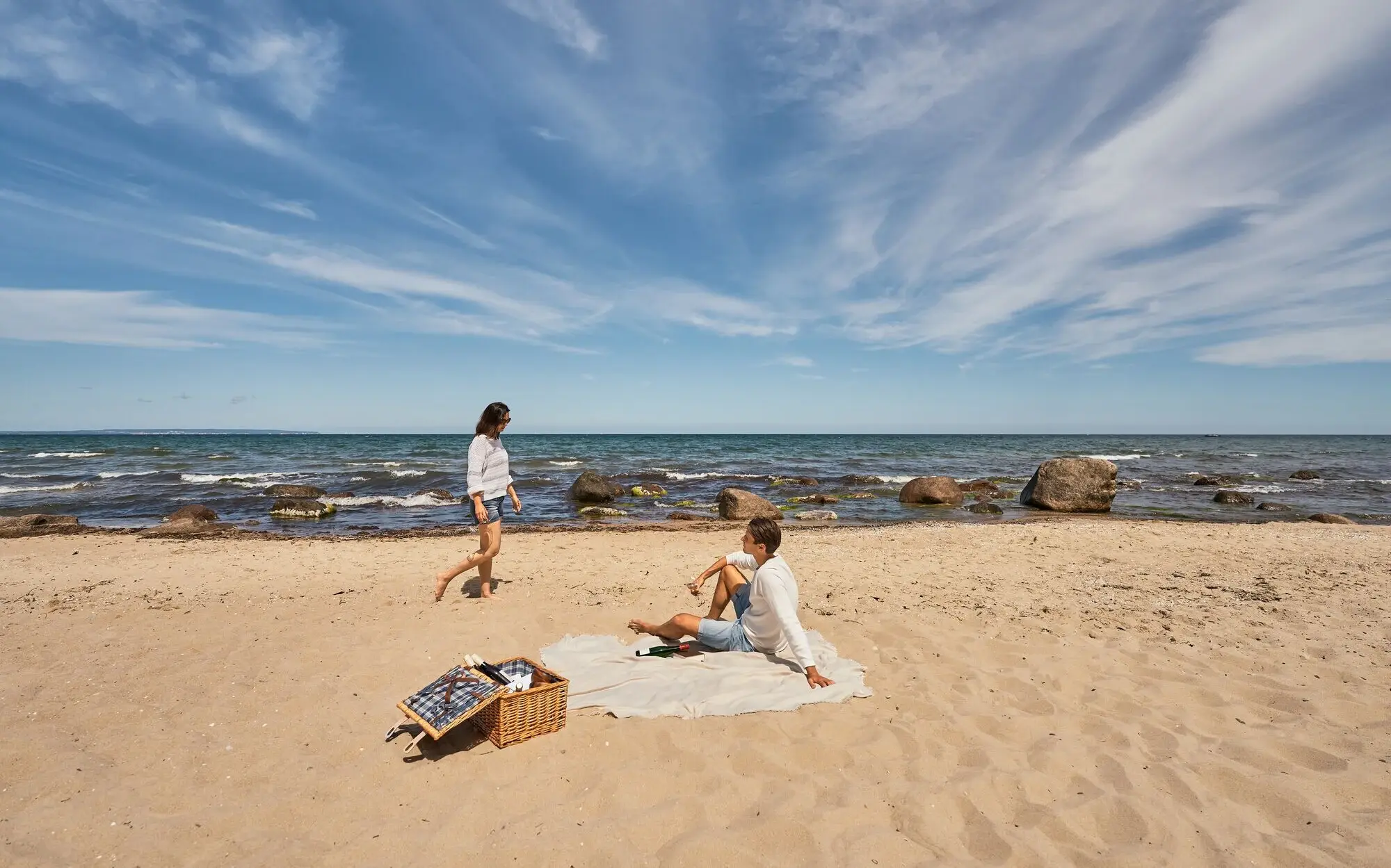 Paar beim Picknick am naturbelassenen Ostseestrand; der Mann sitzt auf einer Decke im Sand, die Frau spaziert barfuß am Wasser entlang, im Vordergrund ein Picknickkorb, im Hintergrund das Meer mit Felsen und weiter Horizont.