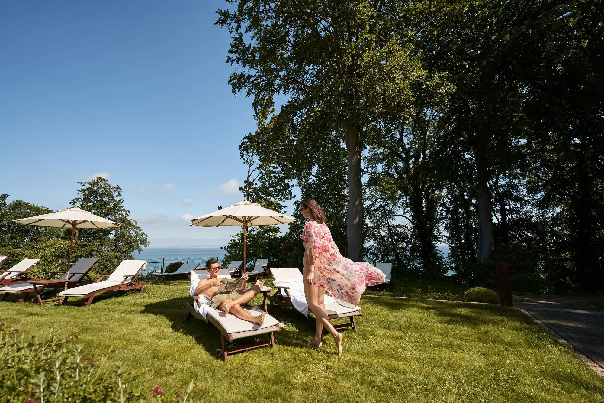 Paar entspannt auf Sonnenliegen im Garten eines Hotels mit Ostseeblick; die Frau trägt ein luftiges Sommerkleid, der Mann prostet mit einem Getränk – Sonnenschirme und grüne Bäume im Hintergrund.