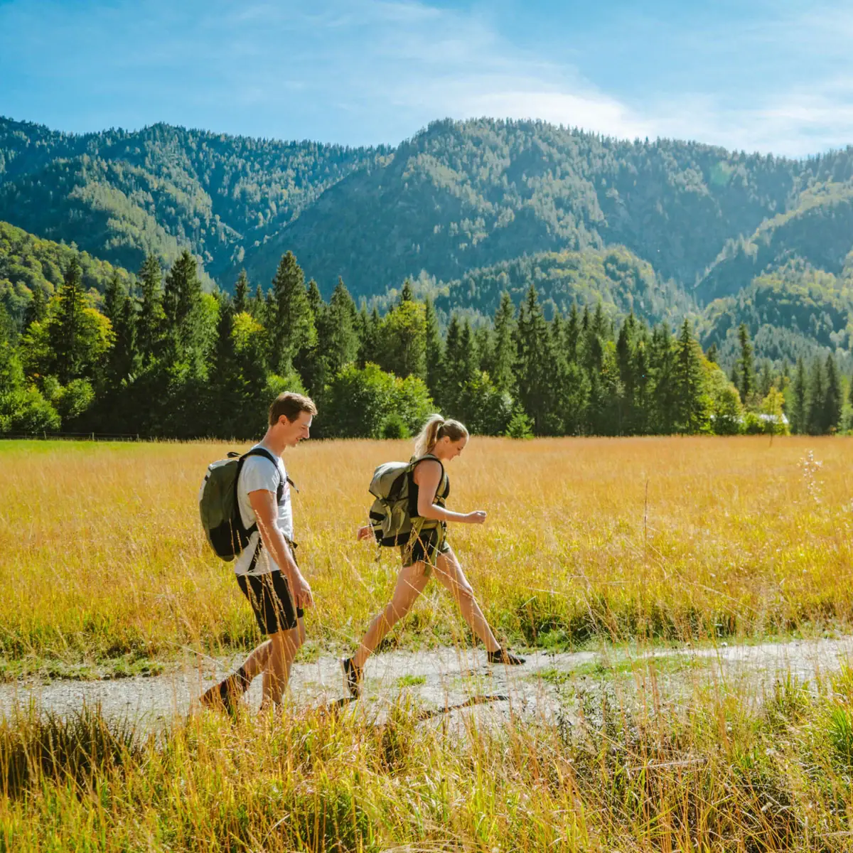 Wanderung im Tal Ein Mann und eine Frau wandern auf einem Pfad in einem Feld mit Bergen im Hintergrund.