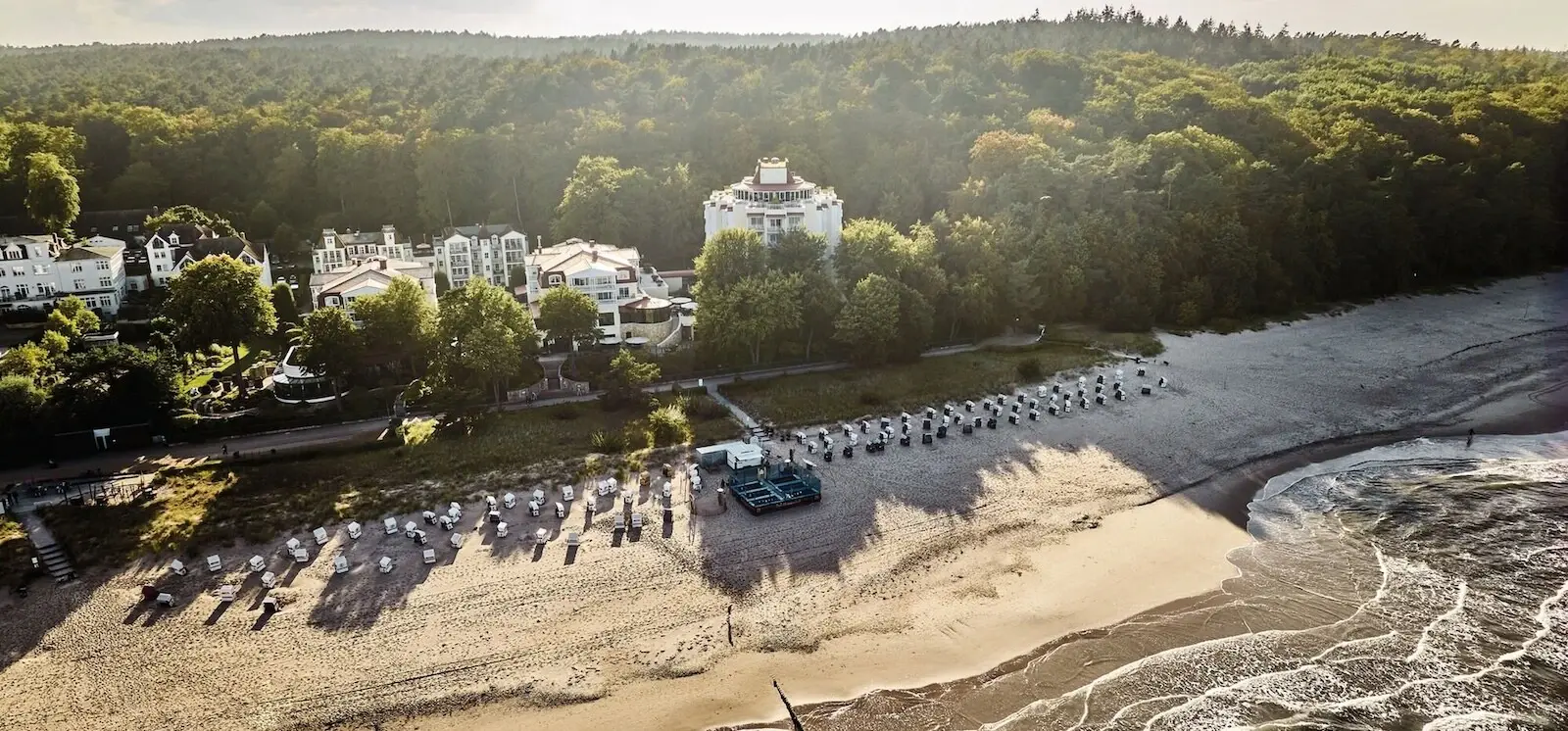 Eine Küstenlandschaft von oben mit vielen Strandkörben am Strand und einigen Häusern zwischen vielen grünen Bäumen.