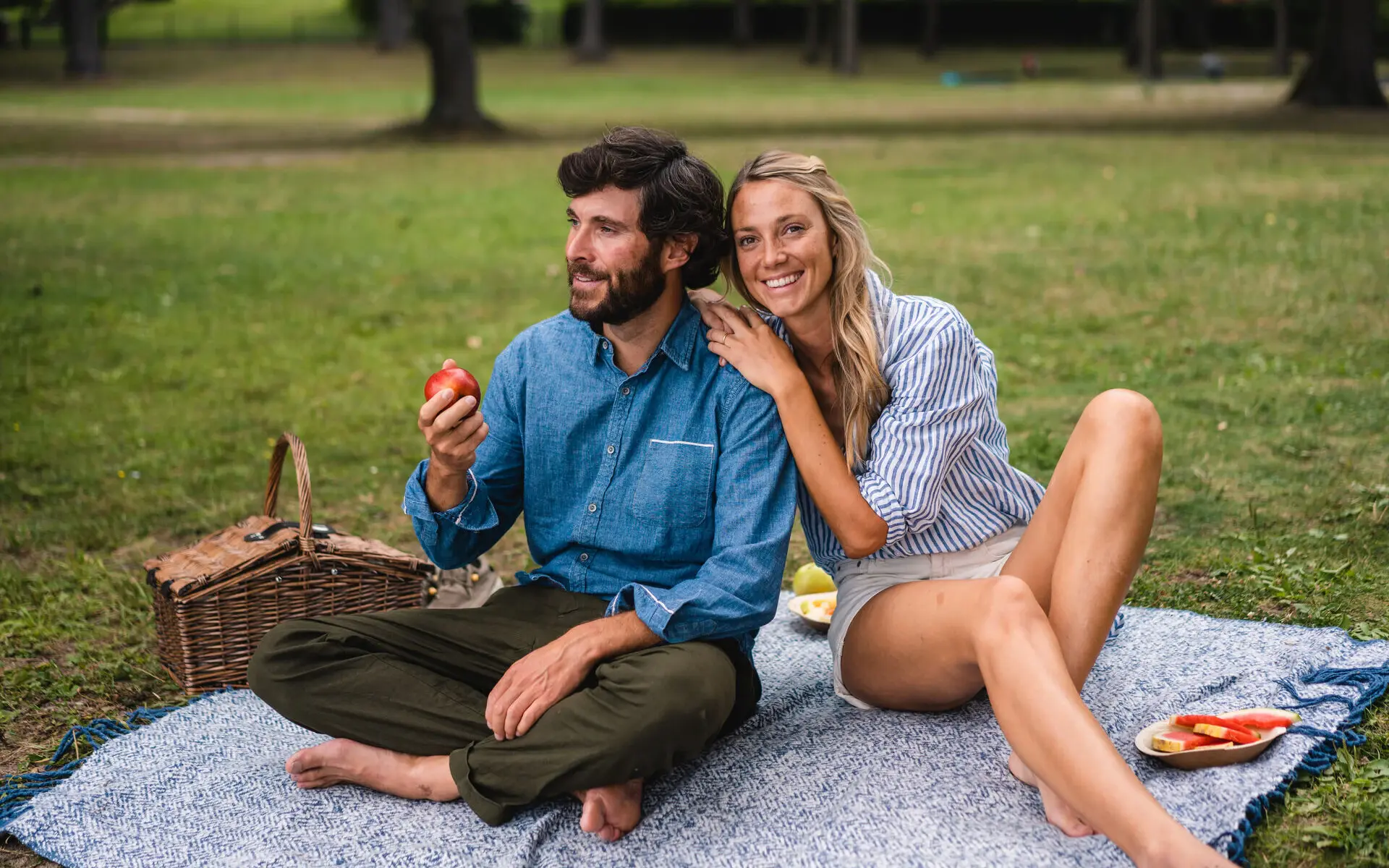 Ein Paar sitzt auf einer Picknickdecke auf einer Wiese und essen Obst. 