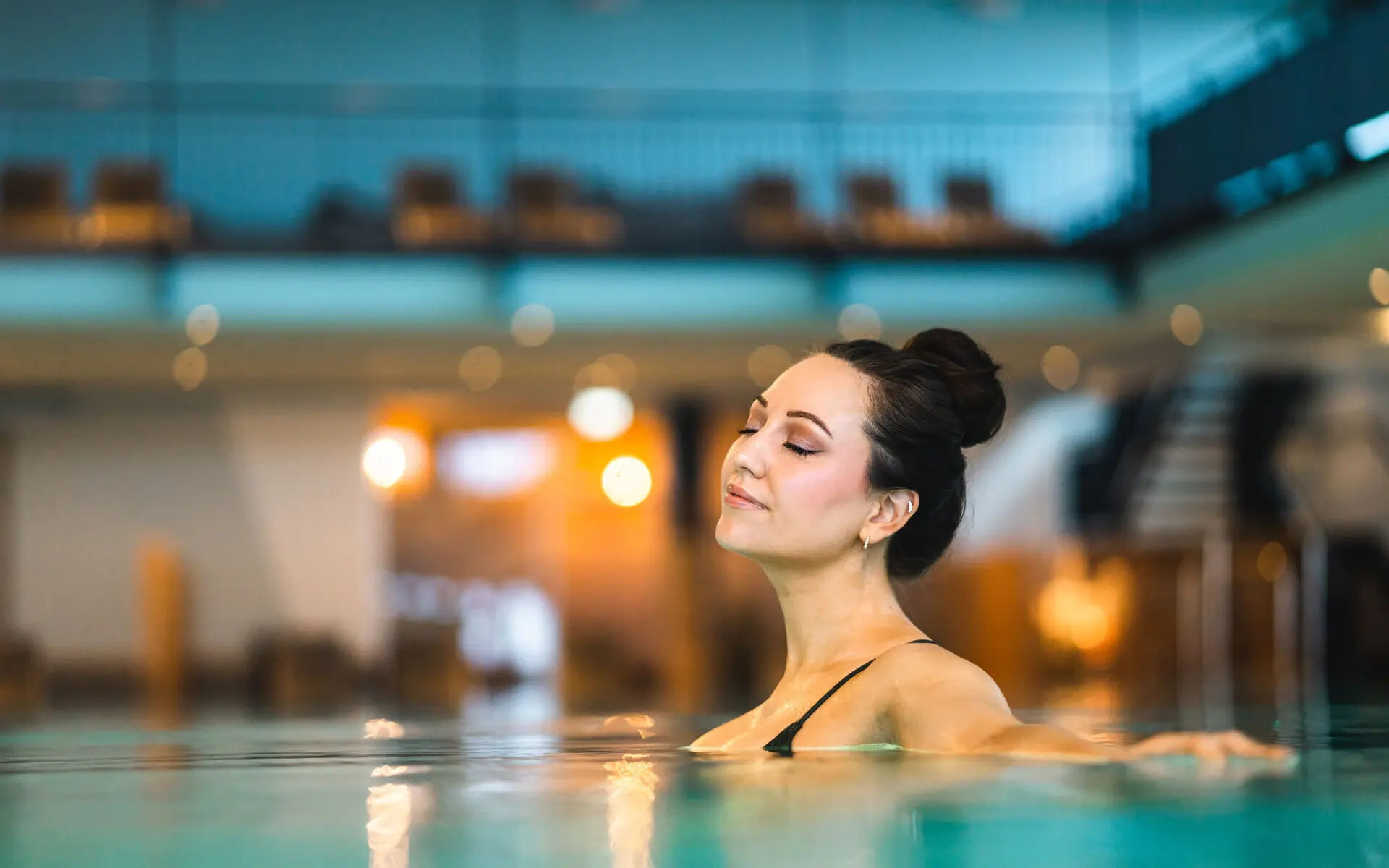 Eine Frau entspannt sich im warmen Wasser eines stilvoll beleuchteten Innenpools. Sie lehnt sich zurück, genießt den Moment mit geschlossenen Augen, während die harmonische Atmosphäre des Wellnessbereichs Ruhe und Gelassenheit ausstrahlt.