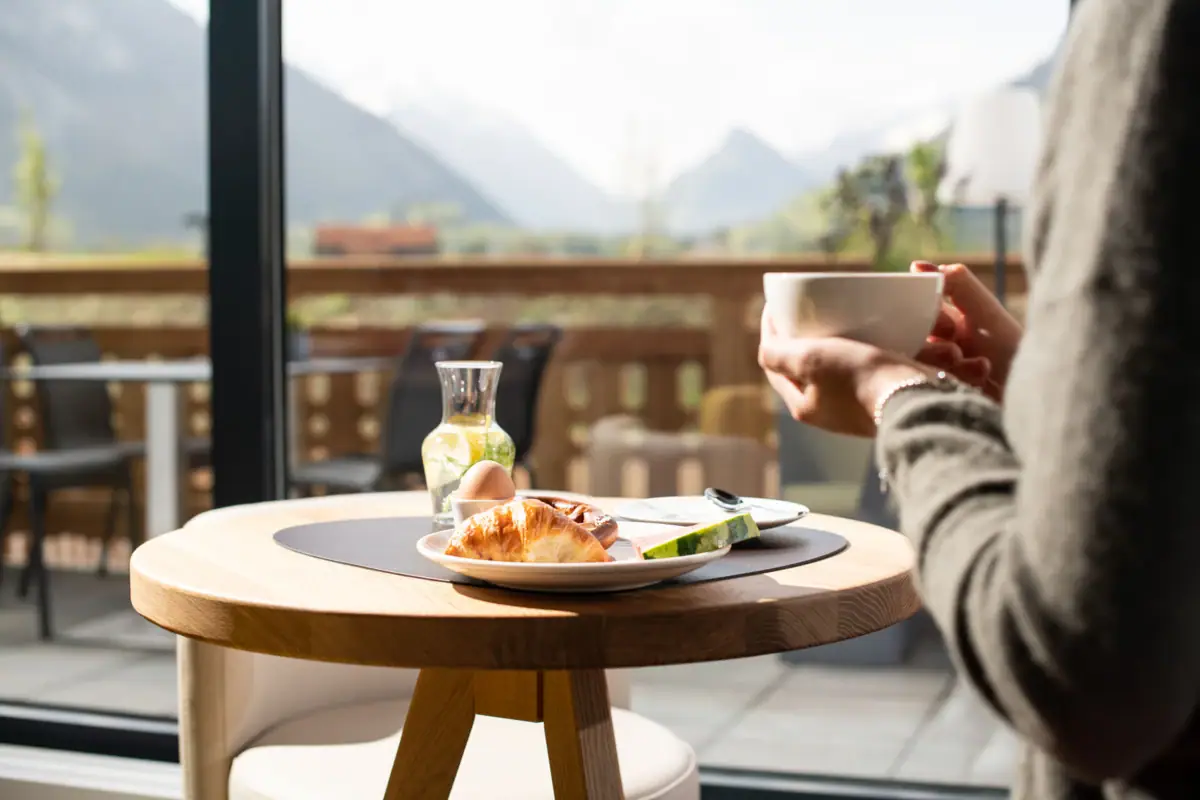 Frühstück mit Bergblick im aja Ruhpolding Eine Person hält eine Tasse Kaffee und einen Teller mit Essen auf einem Tisch.