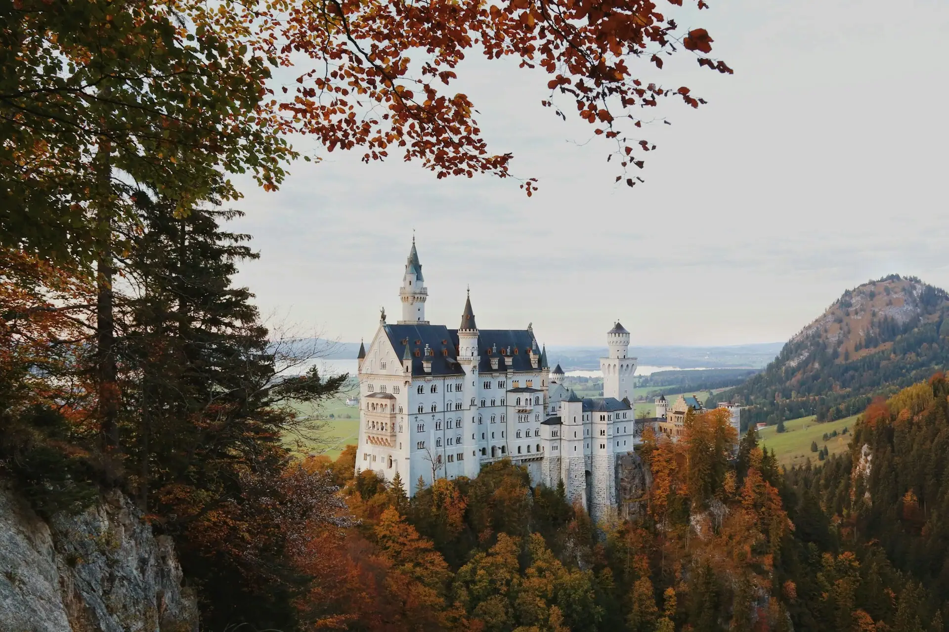 Schloss Neuschwanstein Ein Schloss auf einem Hügel mit Bäumen und Bergen im Hintergrund.