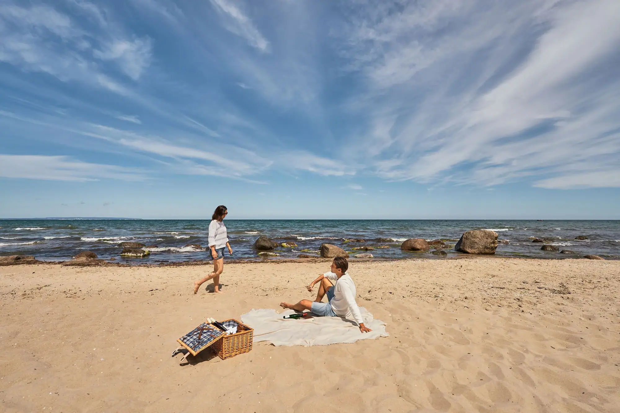 Paar beim Picknick am naturbelassenen Ostseestrand; der Mann sitzt auf einer Decke im Sand, die Frau spaziert barfuß am Wasser entlang, im Vordergrund ein Picknickkorb, im Hintergrund das Meer mit Felsen und weiter Horizont.