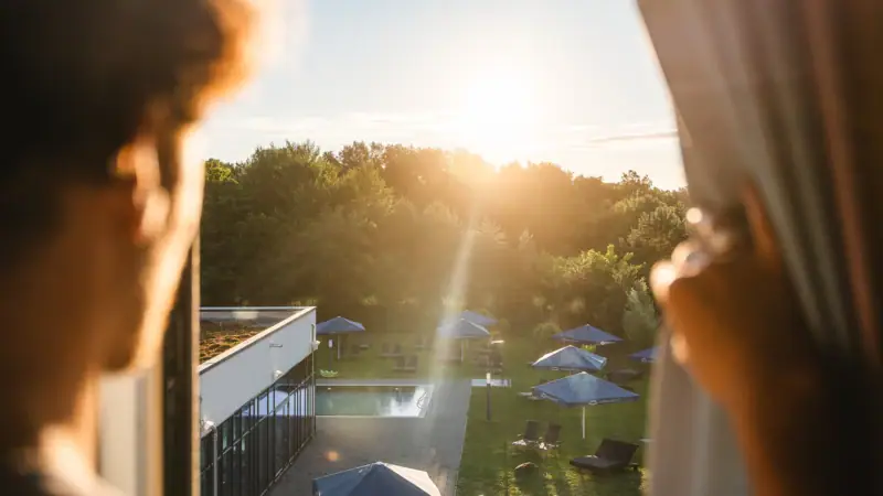 Zimmer im aja Bad Saarow Eine Person betrachtet einen Pool mit Sonnenschirmen und Bäumen.