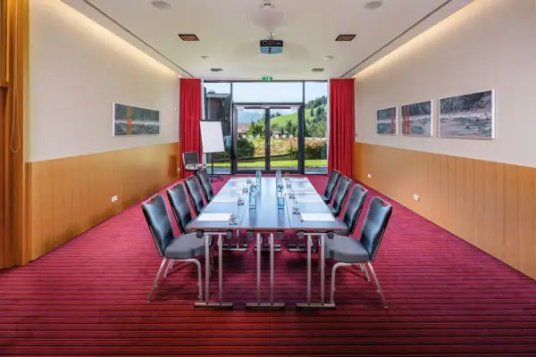 In einem Tagungsraum mit rotem Teppich steht ein langer Tisch. Durch eine große Terrassentür im Hintergrund fällt Tageslicht herein. 