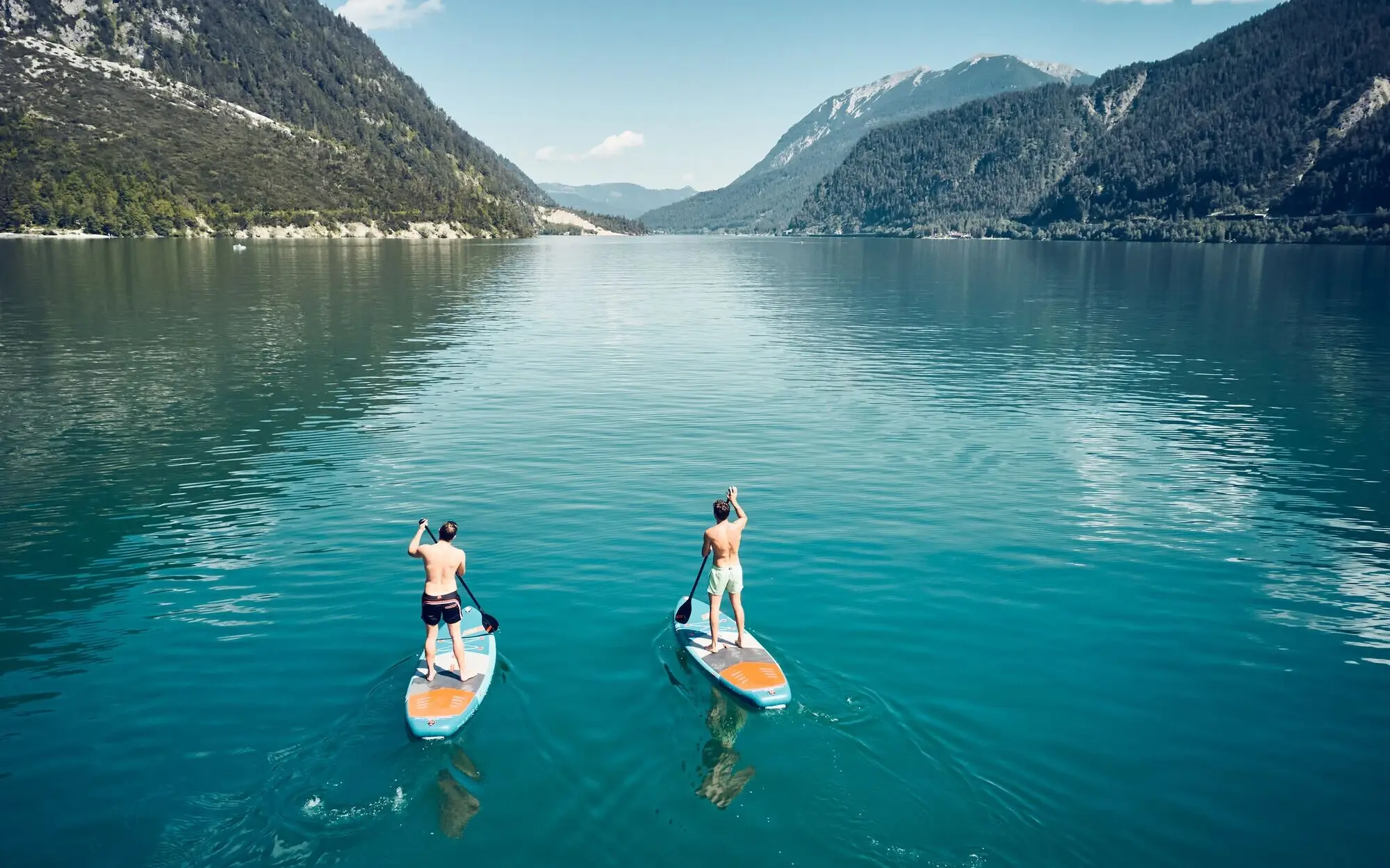 Zwei Männer auf Paddleboards auf einem See mit Bergen im Hintergrund.