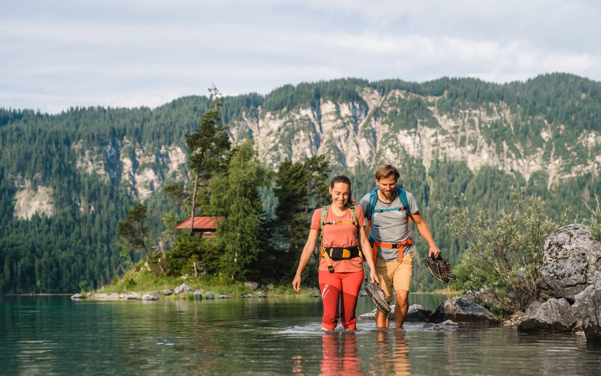 Ein Paar in Wanderkleidung spaziert durch das flache Wasser eines Sees. Im Hintergrund erstrecken sich die Weiten der Berge. 