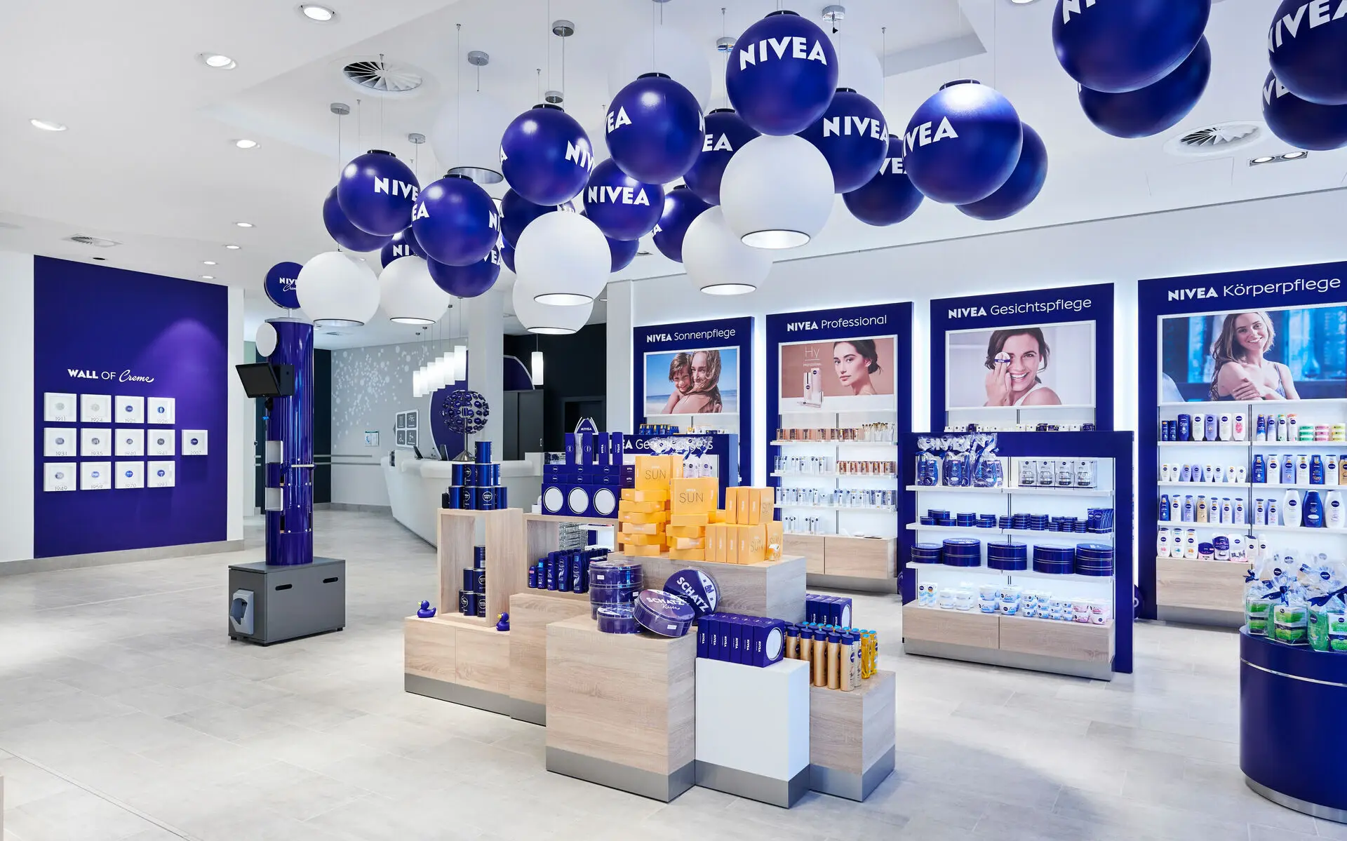 In einem Verkaufsraum mit hellem Holzboden und weißen und dunkelblauen Wänden sind diverse NIVEA Produkte in den Regalen und verschiedenen Verkaufsaufstellern zu erkennen. An der Decke hängen blaue und weiße Luftballons mit dem NIVEA Logo bedruckt.