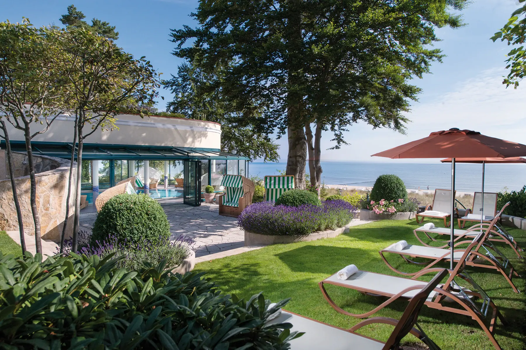 Liegestühle stehen draußen auf einer Wiese mit Meerblick und im Hintergrund ist ein Indoor-Pool zu sehen.