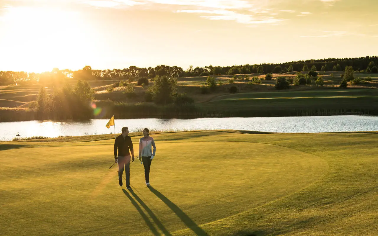 Zwei Personen mit Golfschlägern in ihren Händen laufen über einen Golfplatz mit Wasser im Hintergrund bei untergehender Sonne. 