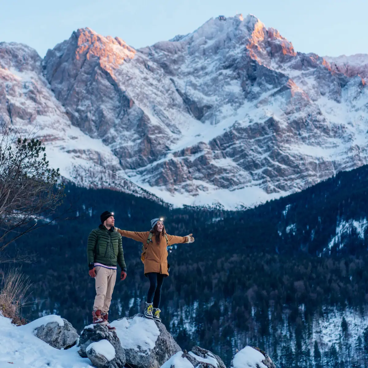 Ein Mann und eine Frau stehen auf einem Felsen mit Schnee und Bergen im Hintergrund.