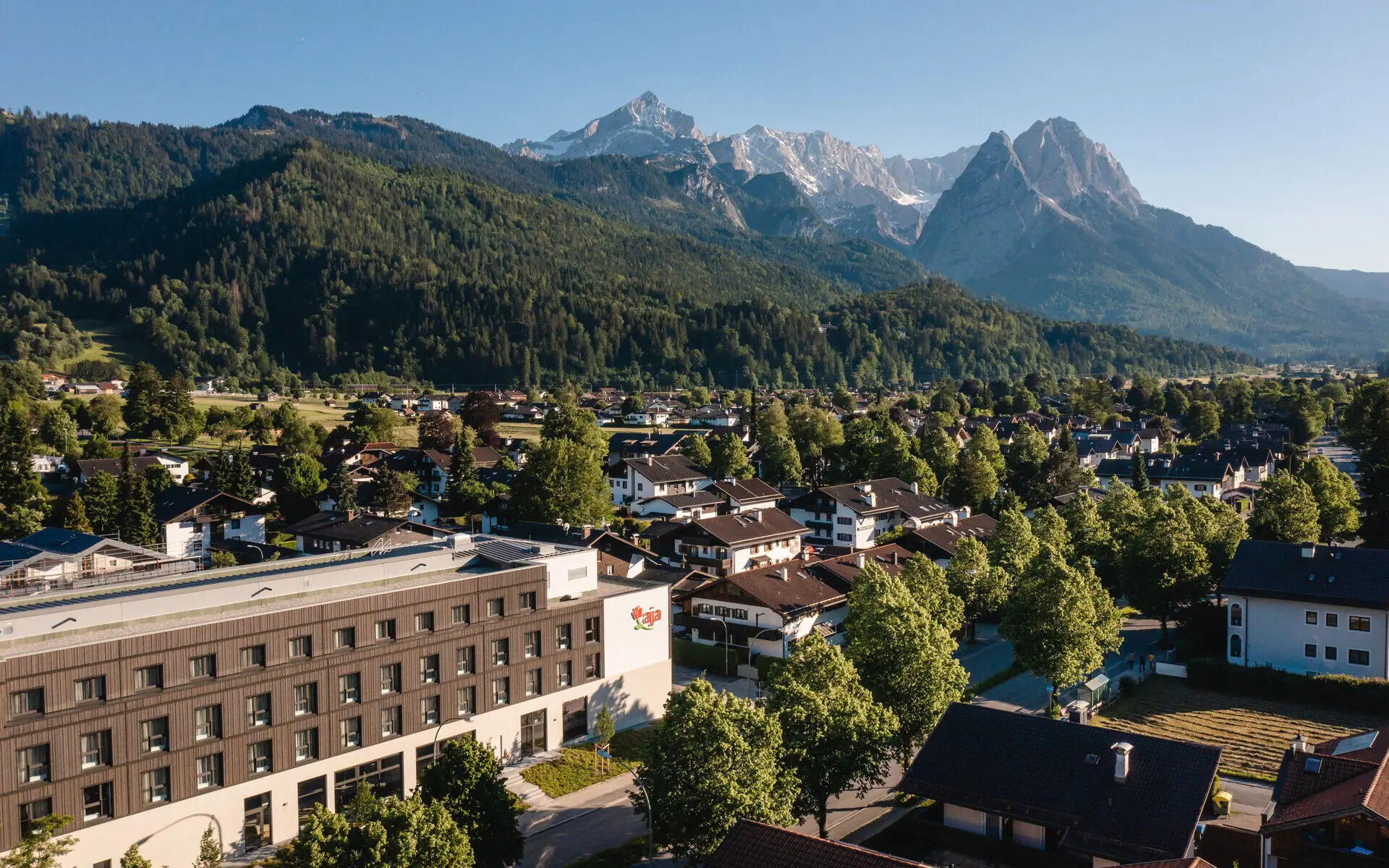 Luftansicht des aja Garmisch-Partenkirchen in einem Dorf und den angrenzenden Bergen im Hintergrund. 