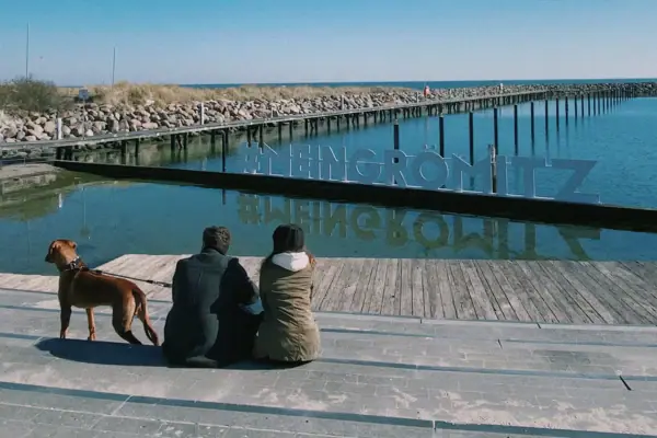 Menschen sitzen auf einem Steg mit einem Hund am Wasser.
