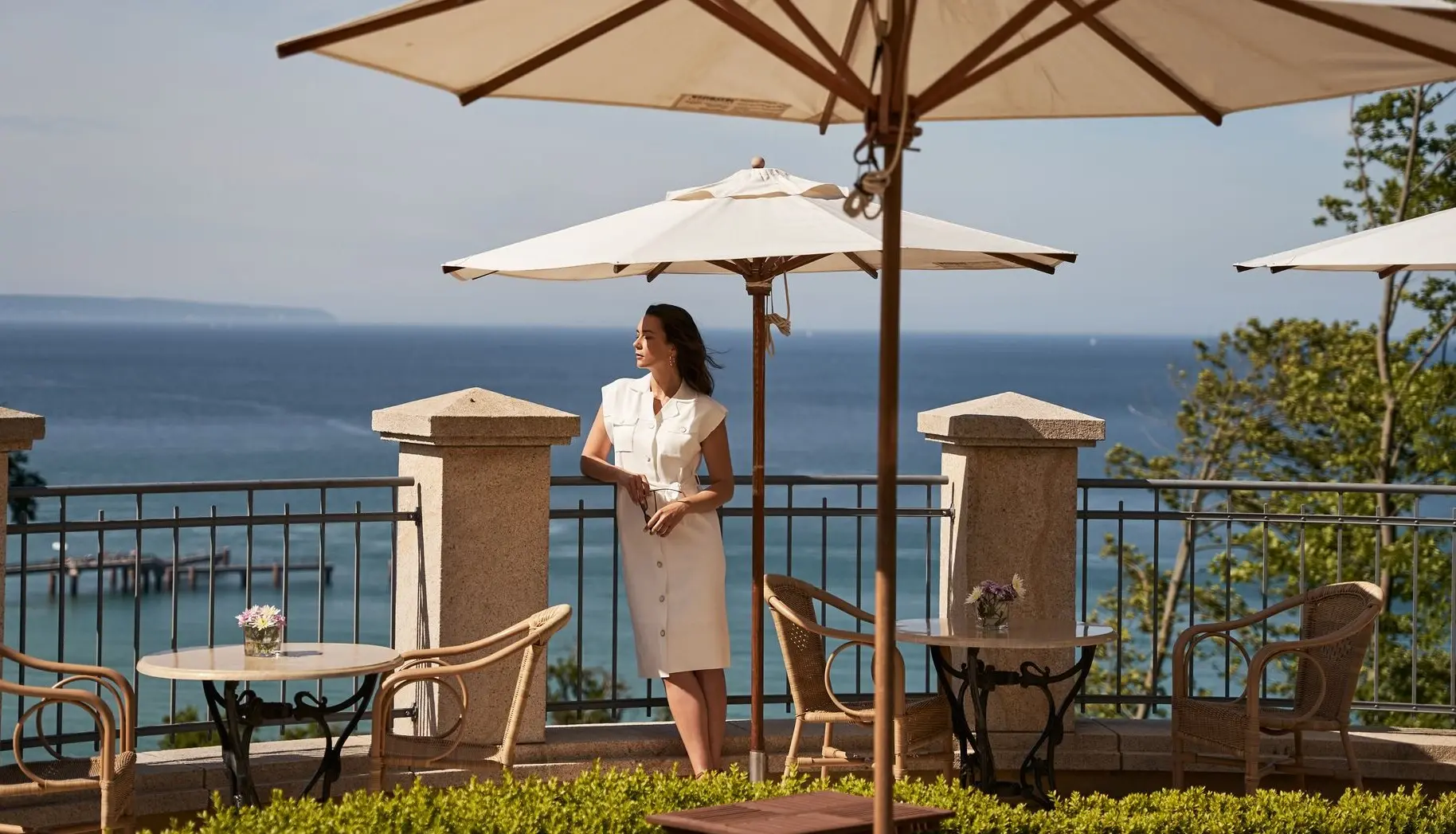 Woman standing under a parasol on a raised outdoor platform with a panoramic view of the sea in the background.