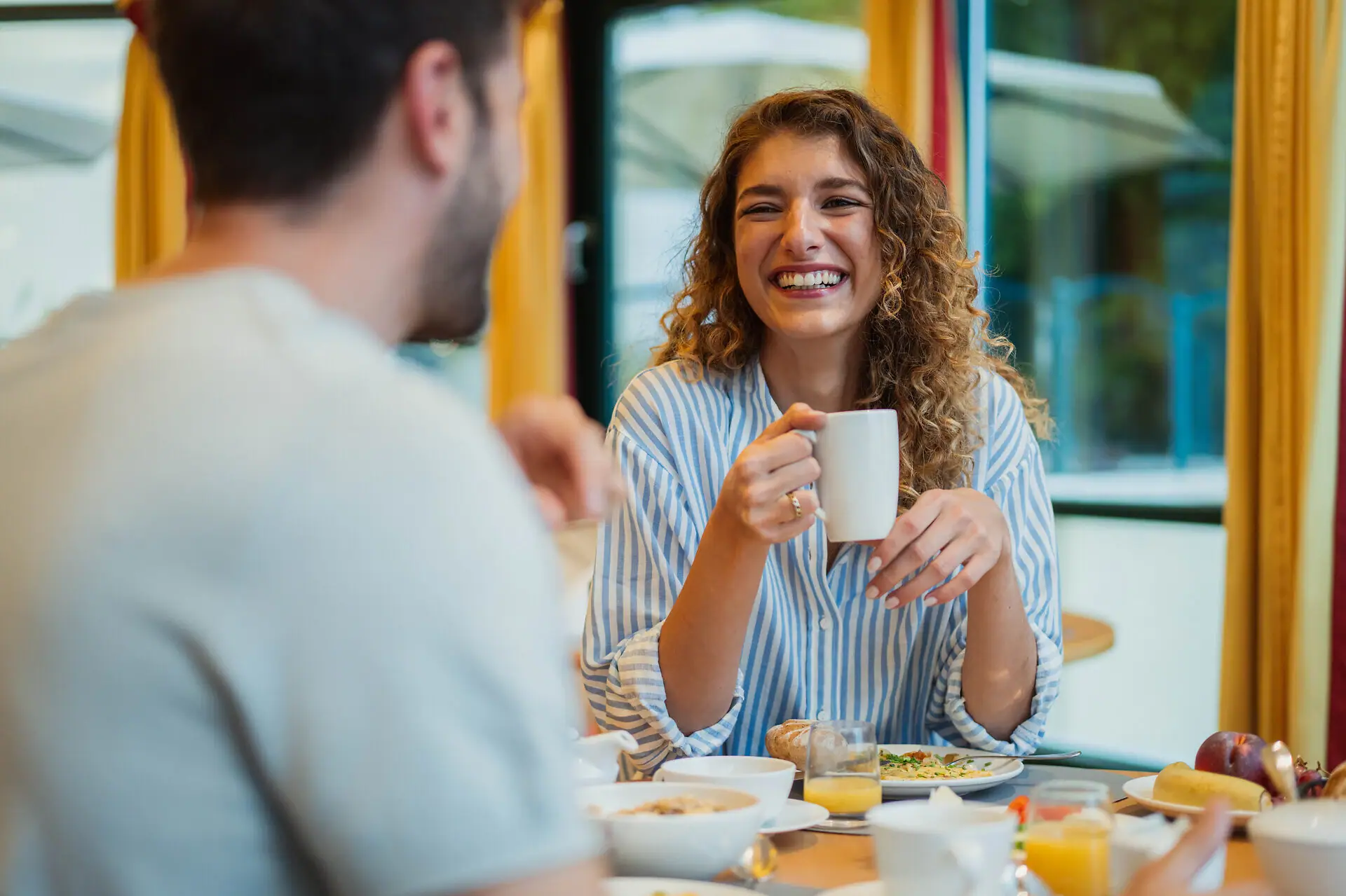 Restaurant Eine Frau und ein Mann sitzen an einem Frühstückstisch in einem Restaurant und die Frau hält lachend eine Tasse in der Hand.