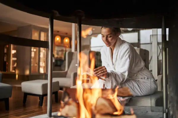 Eine Frau in einem weißen Bademantel sitzt auf einer Steinfläche am Feuer.
