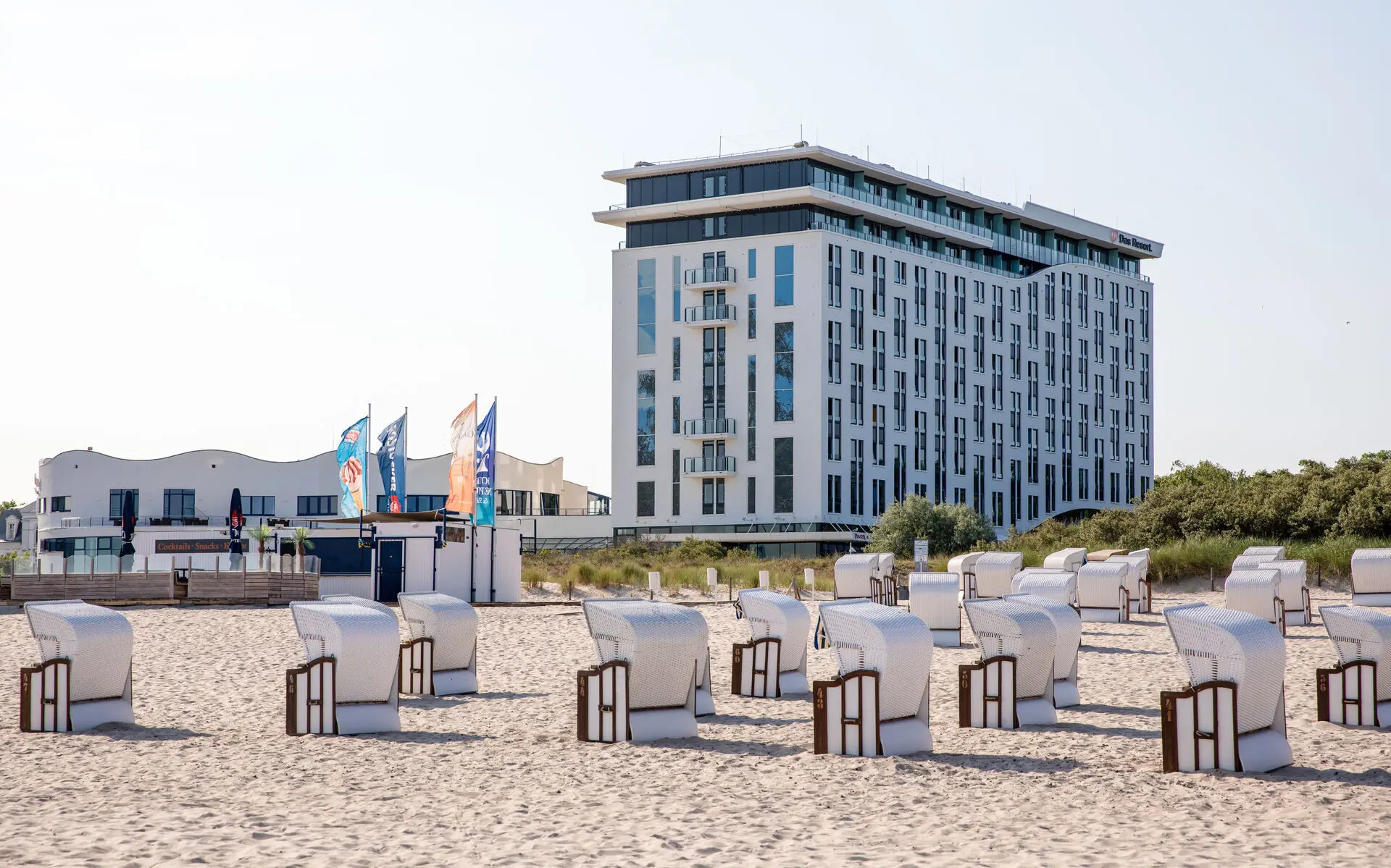 Vor einem weißen Gebäude am Strand stehen Strandkörbe aufgereiht und die Sonne scheint. 