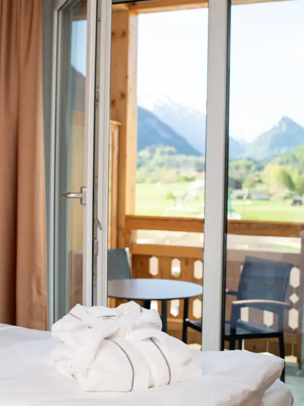 Zimmer aja Ruhpolding Ein weißer Bademantel liegt auf einem Bett vor einem geöffneten Balkonfenster mit Blick auf die Berge.