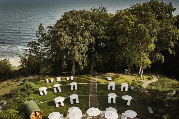 Eine Gruppe weißer Sonnenschirme auf einer grasbewachsenen Fläche mit Bäumen und dem Meer im Hintergrund.