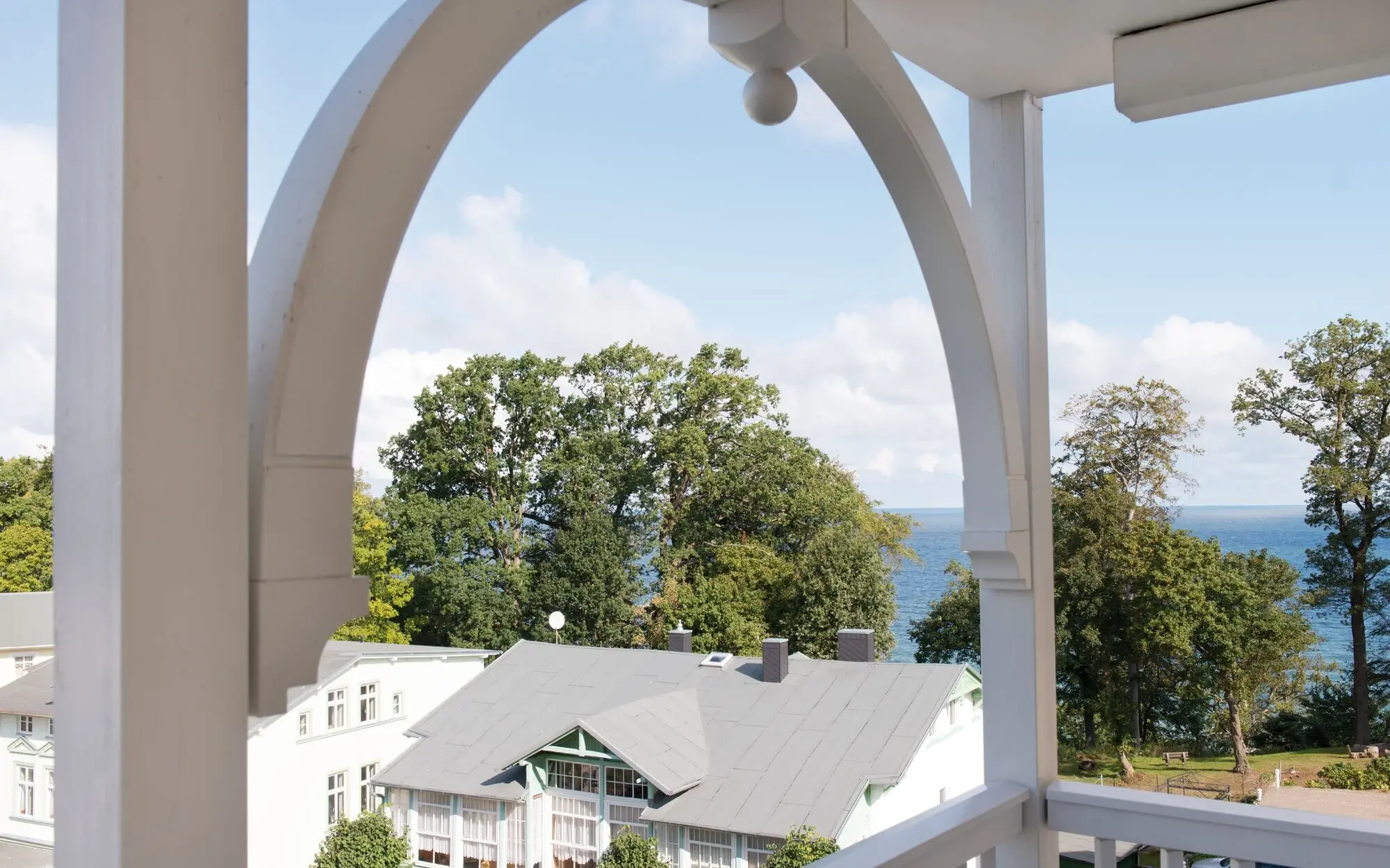 Blick von einem Balkon auf ein Haus mit blauem Himmel und dem Meer im Hintergrund.