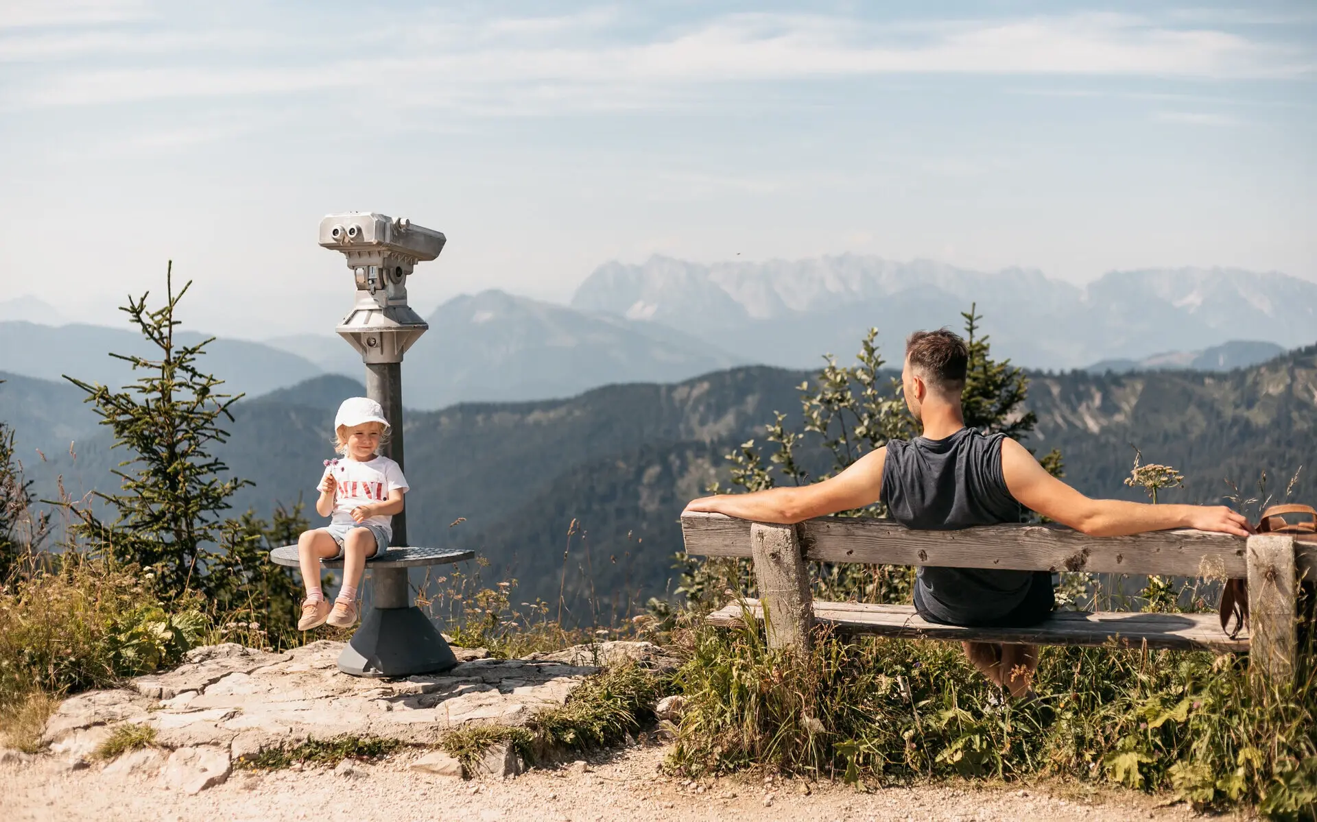 Ein Mann sitzt auf einer Holzbank, neben der ein Fernglas steht, auf dem ein kleines Mädchen sitzt, zu dem er hinüber schaut. Im Hintergrund erstrecken sich die Weiten der Berge und die Sonne scheint warm vom Himmel.