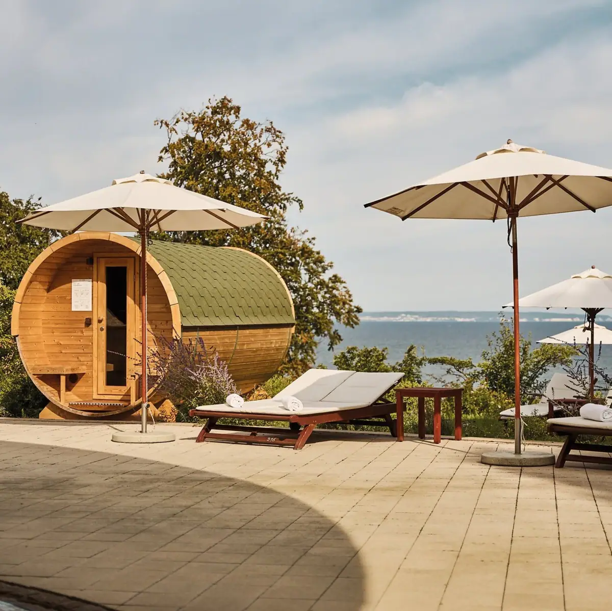 Faßsauna mit Meerblick Liegestühle und Sonnenschirme neben einem Fass und einem Gewässer.